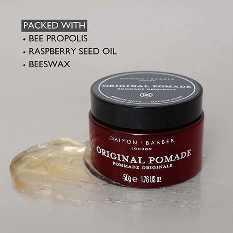 A red jar of Daimon Barber Original Pomade with a black lid and a scoop of translucent product next to it, with text on the jar and lid reading Daimon Barber London Original Pomade Pommade Originale, 50g e 1.76 US oz, Medium Hold, Medium Shine, and a list of ingredients in the upper left corner stating PACKED WITH Bee Propolis Raspberry Seed Oil Beeswax.