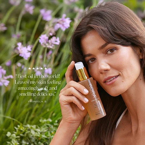 Image 1 - A woman with long dark hair holds a bottle of oil and smiles slightly, surrounded by purple flowers and greenery, with text that reads Best oil in the world! Leaves my skin feeling amazingly soft and smelling delicious. GABRIELA D.
              Image 2 - A hand holding a bottle of KORA Organics Noni Glow Body Oil against a bright blue sky, with visible text that reads Best oil in the world! Leaves my skin feeling amazingly soft and smelling delicious. Gabriela D.
              Image 3 - Image of two body oil products from KORA Organics, with a new packaging design on the right; the left bottle is yellow with a pump top, and the right is a tan bottle, both labeled with KORA ORGANICS and NON-GLOW BODY OIL, alongside floral illustrations and the text New Look, Same Formula.