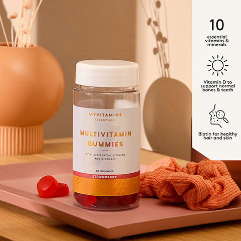 A bottle of Myvitamins Essentials Multivitamin Gummies in Strawberry flavor is shown on a pink tray with two gummies and a scrunchie alongside text highlighting benefits MYVITAMINS ESSENTIALS MULTIVITAMIN GUMMIES WITH 10 ESSENTIAL VITAMINS AND MINERALS 30 GUMMIES STRAWBERRY 10 essential vitamins & minerals Vitamin D to support normal bones & teeth Biotin for healthy hair and skin.