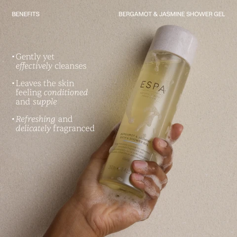 A persons hand, covered in white soap suds, holds a clear bottle of yellow shower gel with a light grey cap against a textured light brown background, with the visible text BENEFITS, Gently yet effectively cleanses, Leaves the skin feeling conditioned and supple, Refreshing and delicately fragranced, BERGAMOT & JASMINE SHOWER GEL, ESPA, NATURAL BEAUTY INNER CALM, BERGAMOT & JASMINE BATH & SHOWER GEL, Soap-free body cleanser for refreshed, delicately fragranced skin, gel nettoyant pour le corps sans savon une peau rafraîchie et délicatement, CLEANSE, 250ml e 8.4 US fl oz.