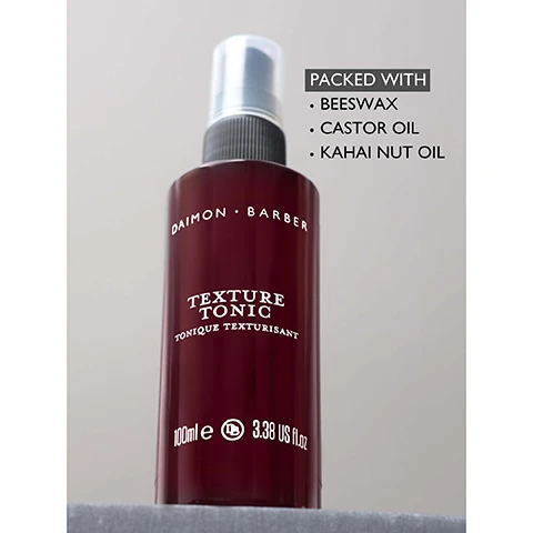 Image 3 - A maroon spray bottle for hair product is displayed, labeled DAIMON · BARBER, TEXTURE TONIC, TONIQUE TEXTURISANT, 100mle e 3.38 US FL.OZ, with an adjacent text box stating PACKED WITH BEESWAX CASTOR OIL KAHAI NUT OIL. Image 4 - A dark red bottle of Daimon Barber Texture Tonic with a black pump dispenser is displayed alongside a customer review that features five stars and reads: Texture Miracle, I have now used this product daily for the past couple of months and have to admit it is the best tonic ever! I get a strong texture and it also makes my hair much easier to manage. The smell is phenomenal and I will be looking to stock up on Daimon Barber products in the next few weeks to come. Highly recommend it to anyone with thick hair. - Happy Customer. The bottle itself is labeled DAIMON BARBER, TEXTURE TONIC, TONIQUE TEXTURISANT, 100ml e, 3.38 US fl. oz.