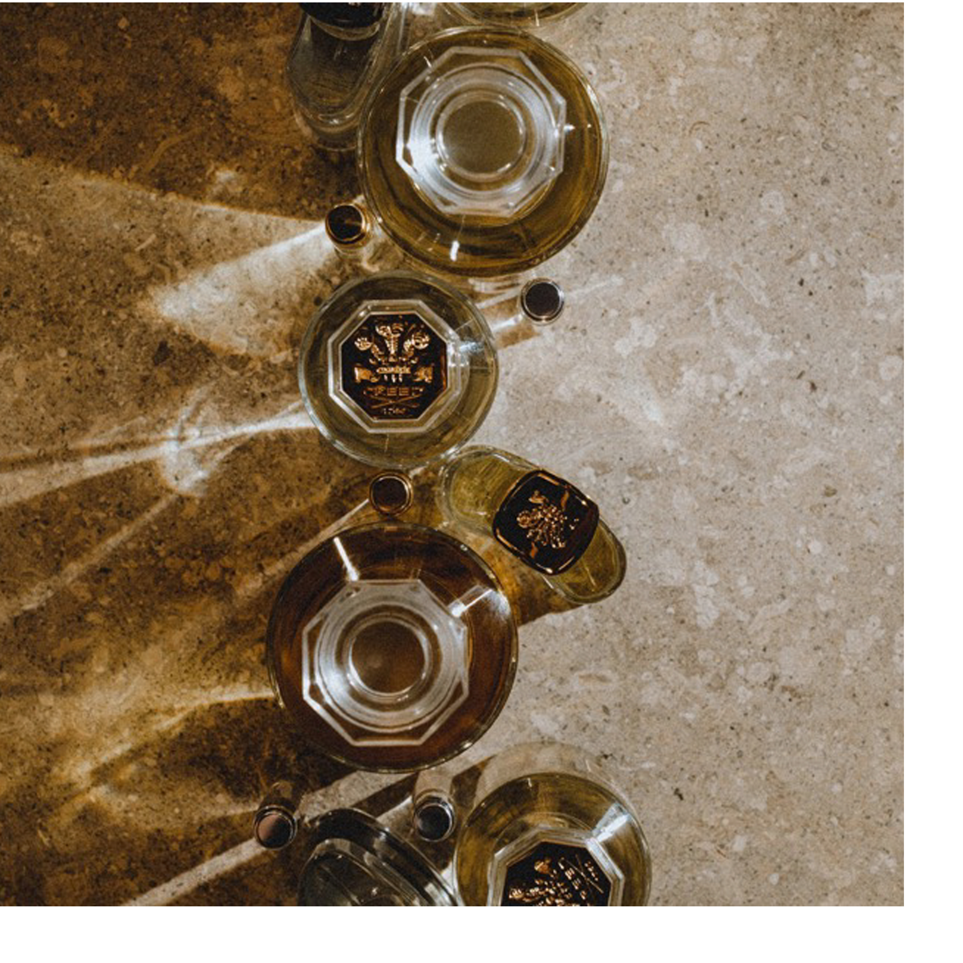 Overhead view of several perfume bottles on a stone surface with diagonal light patterns, one bottle shows the text CREED 1760.