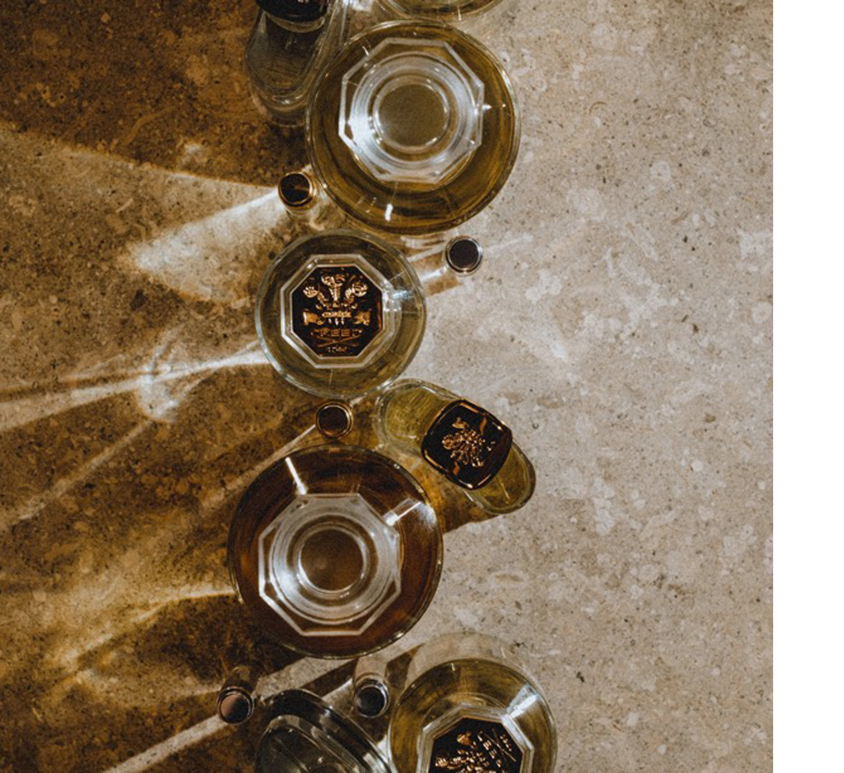 Overhead view of several perfume bottles on a stone surface with diagonal light patterns, one bottle shows the text CREED 1760.