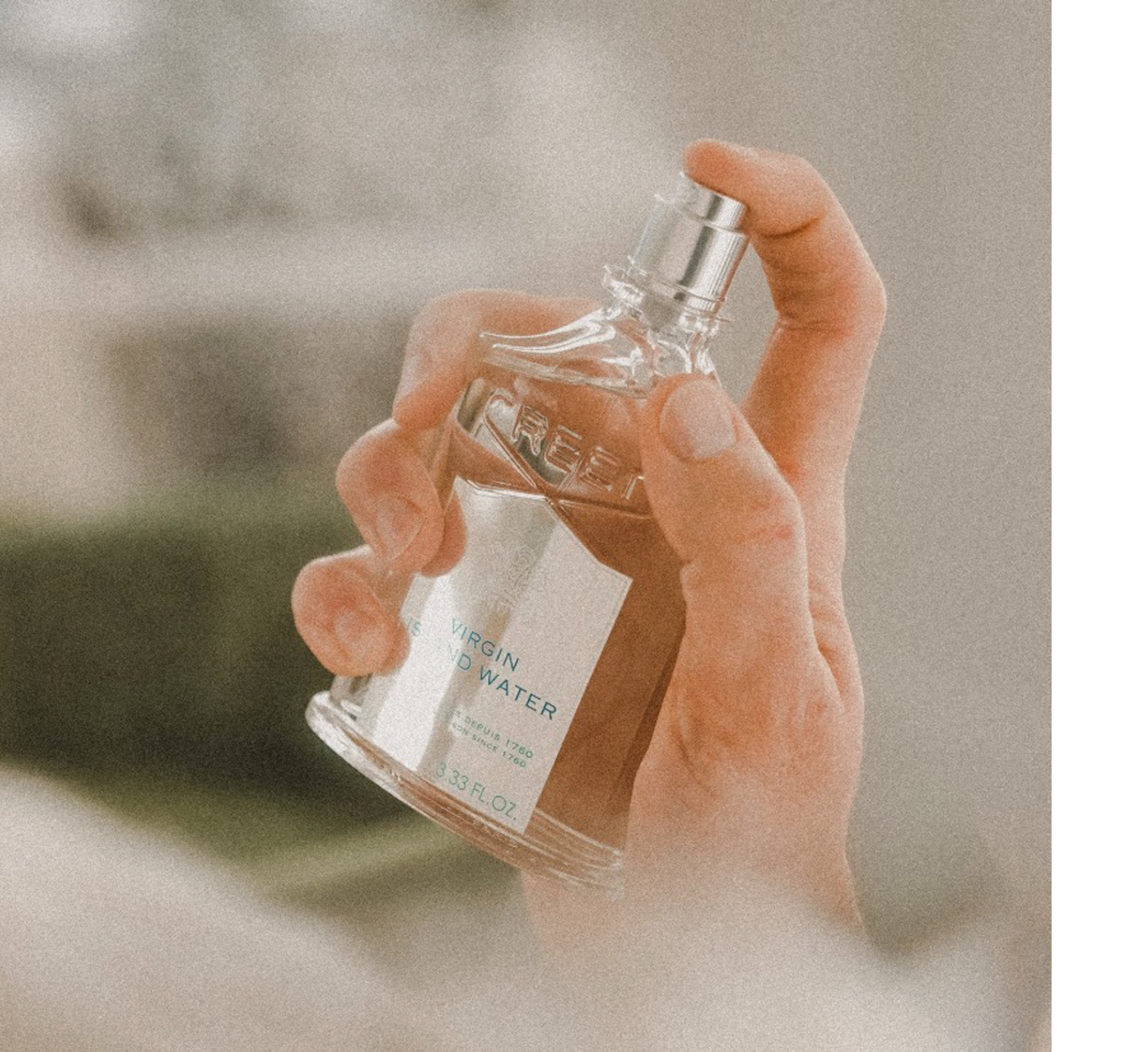 A hand holds a glass bottle of Creed Virgin Island Water perfume showing the label with text VIRGIN ISLAND WATER, FRANCAIS DEPUIS 1760 ENGLISH SINCE 1760, and 3.33 FL. OZ.
