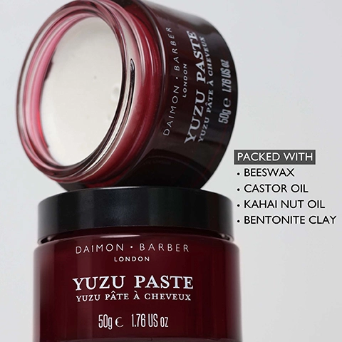 A dark red jar of hair paste with its lid removed is stacked on top of another identical lidded jar, both labeled Daimon Barber London Yuzu Paste Yuzu Pâte à Cheveux 50g e 1.76 US oz, with a list of ingredients to the right stating PACKED WITH Beeswax Castor Oil Kahai Nut Oil Bentonite Clay.