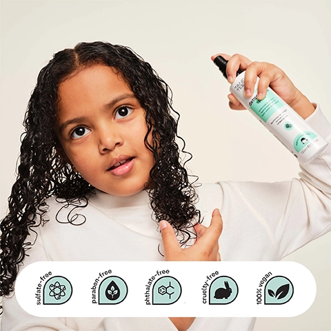 A young child with dark, wet curly hair holds a white spray bottle by Tangle Teezer to their head, while text on the bottle reads detangling spray, moisturizes & protects against breakage for faster, easier detangling, nourishing, lightweight formula with coconut scent, PERFECT FOR KIDS, 150ml e/5 fl.oz, and text on five icons below indicates sulfate-free, paraben-free, phthalate-free, cruelty-free, 100% vegan.