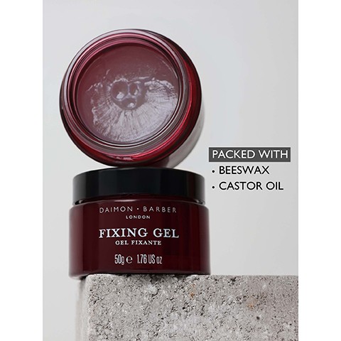 Image 4 - A dark red jar of hair gel sits on a concrete block with an open jar behind it, displaying text Daimon Barber London Fixing Gel Gel Fixante 50g e 1.76 US oz, and an overlay text box states Packed With, Beeswax, Castor Oil.