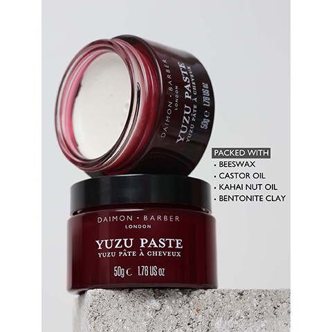 Image 3 - Two dark red jars of Daimon Barber Yuzu Paste are stacked on a concrete block, with the top jar open revealing white paste, and a text box next to them lists ingredients; the visible text on the jars and box includes Daimon Barber London Yuzu Paste Yuzu Pâte à Cheveux 50g e 1.76 US oz and Packed with Beeswax Castor Oil Kahai Nut Oil Bentonite Clay.