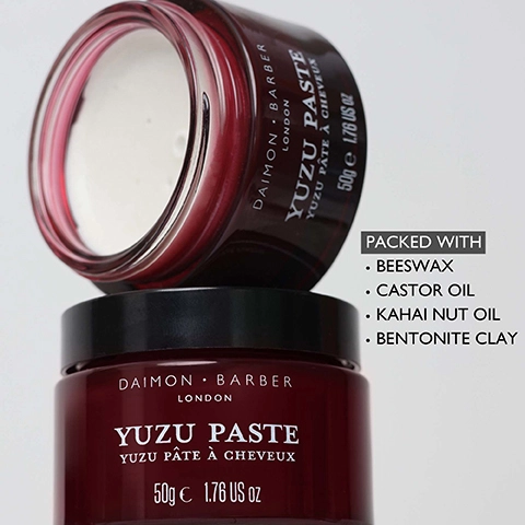 A deep red jar of Daimon Barber London Yuzu Paste, Yuzu Pâte à Cheveux, 50g e 1.76 US oz, sits with an open identical jar stacked on top revealing white paste, and a text box to the right lists PACKED WITH Beeswax, Castor Oil, Kahai Nut Oil, Bentonite Clay.