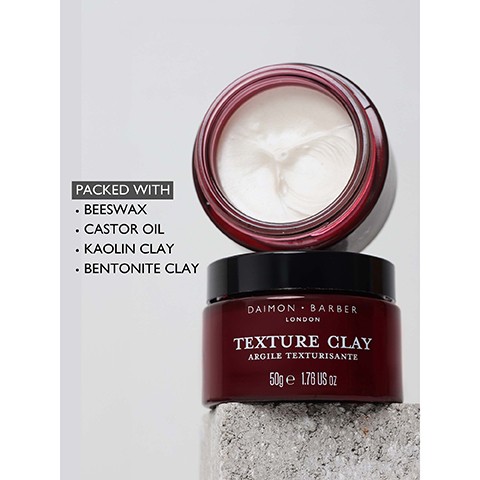 Image 3 - Two dark red jars of hair clay, one open with white product visible and the other closed, are stacked on a concrete block with visible text reading PACKED WITH BEESWAX CASTOR OIL KAOLIN CLAY BENTONITE CLAY and on the jar DAIMON BARBER LONDON TEXTURE CLAY ARGILE TEXTURISANTE 50g e 1.76 US oz.