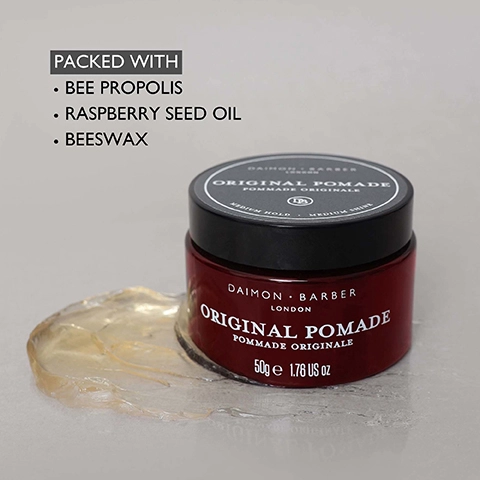 A dark red jar of Daimon Barber Original Pomade with a black lid, showing some clear yellowish pomade spilled on a grey surface, features visible text including PACKED WITH BEE PROPOLIS, RASPBERRY SEED OIL, BEESWAX, DAIMON BARBER LONDON ORIGINAL POMADE POMMADE ORIGINALE, MEDIUM HOLD, MEDIUM SHINE, and 50g e 1.76 US oz.