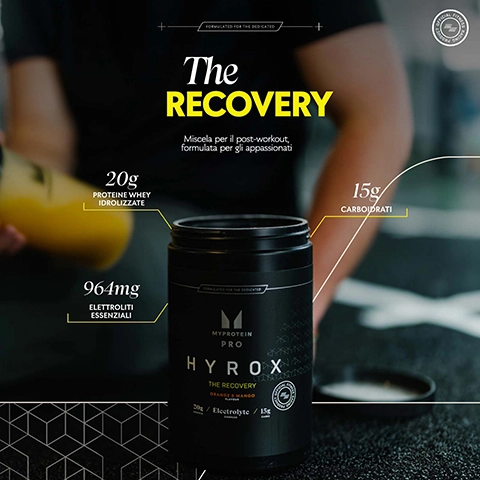 Image of a black protein powder container labeled MYPROTEIN PRO HYROX THE RECOVERY with visible text listing 20g of protein, 15g of carbohydrates, and 964mg of electrolytes; a person’s hand is holding a yellow shaker in the background.