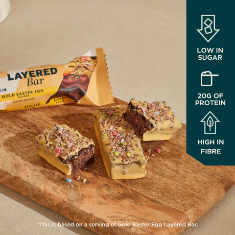 An assortment of chocolate layered protein bars with a creamy coating and sprinkles on a wooden board, alongside a partially visible wrapper labeled Layered Bar with Gold Easter Egg flavour and various nutritional claims including low in sugar, 20g of protein, and high in fibre.