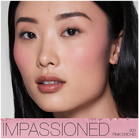 A close-up portrait of a model with long, straight black hair, wearing soft makeup featuring rosy cheeks and glossy lips, with the text that reads IMPASSIONED 903 PINK ORCHID.