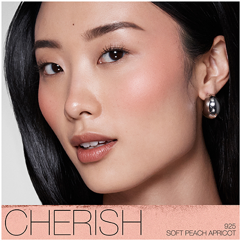 A close-up of a young woman with long black hair and natural makeup, showcasing a soft peach blush, wearing a silver hoop earring, with the text CHERISH 925 SOFT PEACH APRICOT displayed at the bottom.