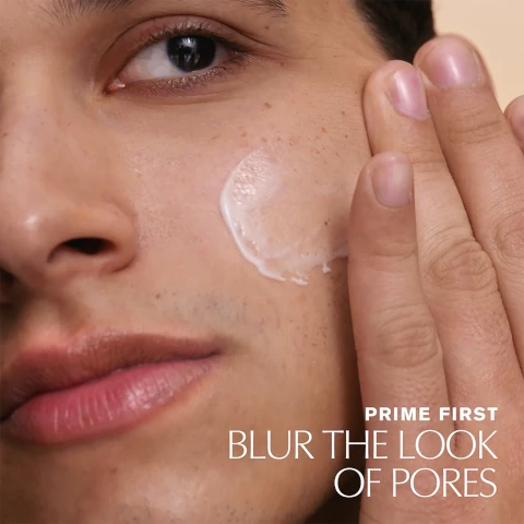 A close-up of a young man applying a cream to his cheek, with the text PRIME FIRST BLUR THE LOOK OF PORES visible at the bottom.