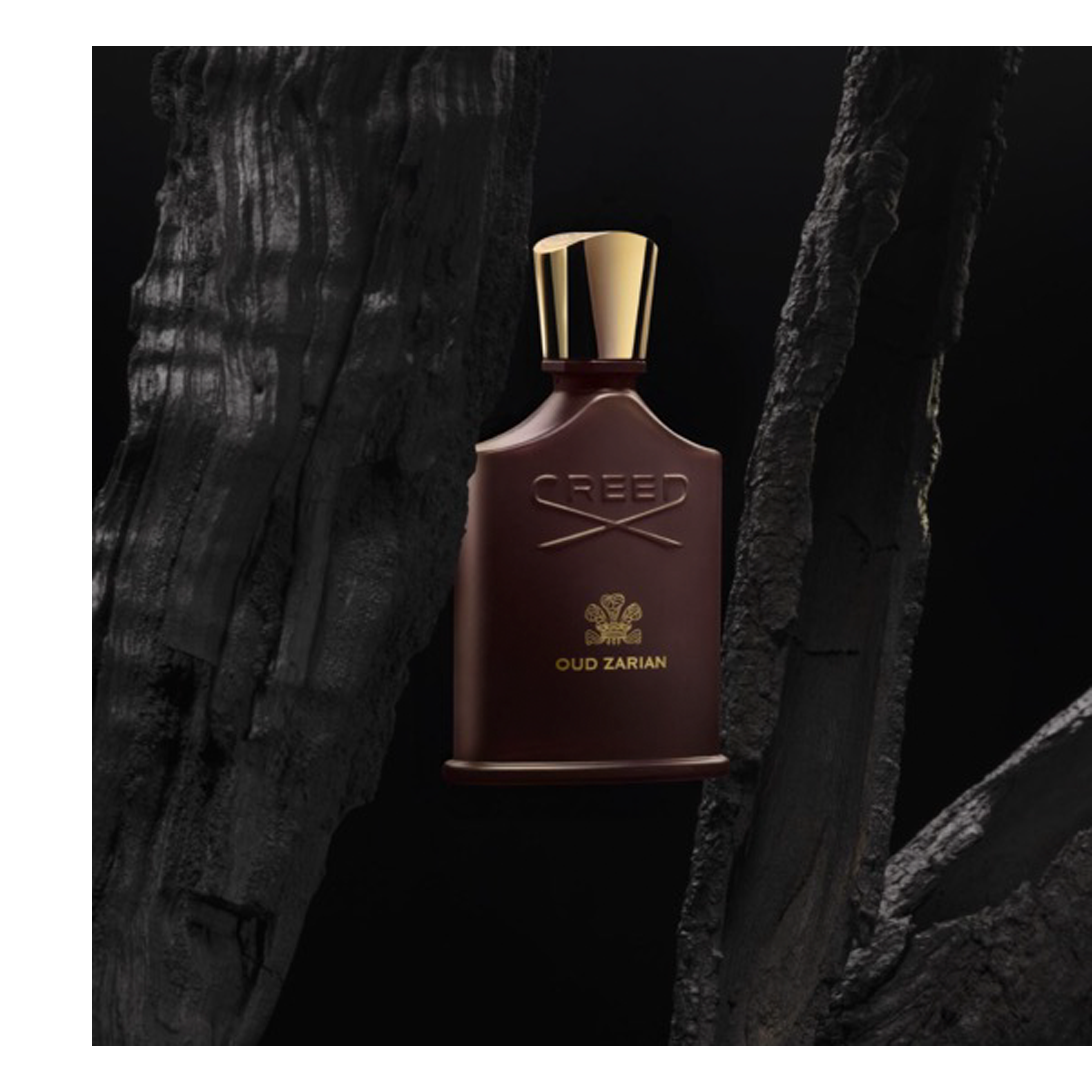 A dark brown perfume bottle with a gold cap and embossed gold text CREED and OUD ZARIAN is centered on a dark background between two dark, rough charred wood pieces.