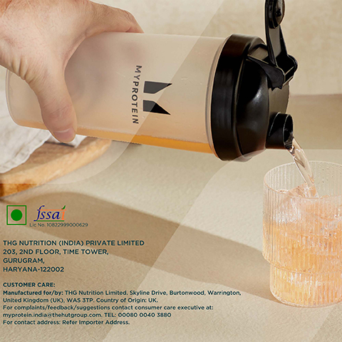 Image 1 - A hand is holding a MyProtein shaker bottle, pouring a light drink into a textured glass on a beige surface, with visible text including FSSAI and manufacturing details.