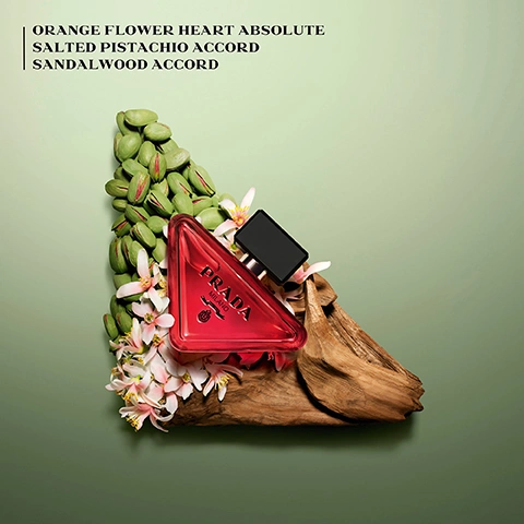 Image 1 - A red triangle perfume bottle sits on driftwood with pistachios and flowers, with text ORANGE FLOWER HEART ABSOLUTE SALTED PISTACHIO ACCORD SANDALWOOD ACCORD PRADA MILANO DAL 1913.