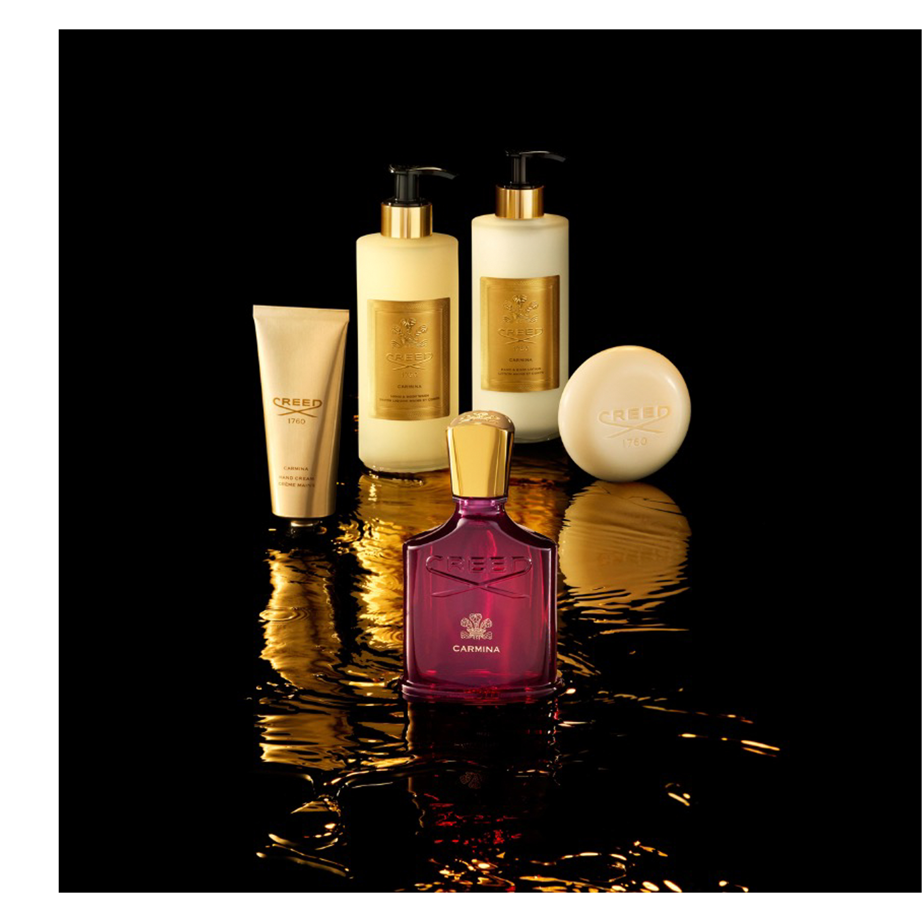 A red Creed Carmina perfume bottle with a gold cap, a beige hand cream tube, two beige pump bottles for hand and body wash and lotion, and a round beige soap bar are displayed on a dark reflective surface with golden liquid effects; visible text on the items includes Creed, 1760, Carmina, Hand Cream, Creme Mains, Hand & Body Wash, Lavant Liquide, Hand & Body Lotion, and Lait Riche.
