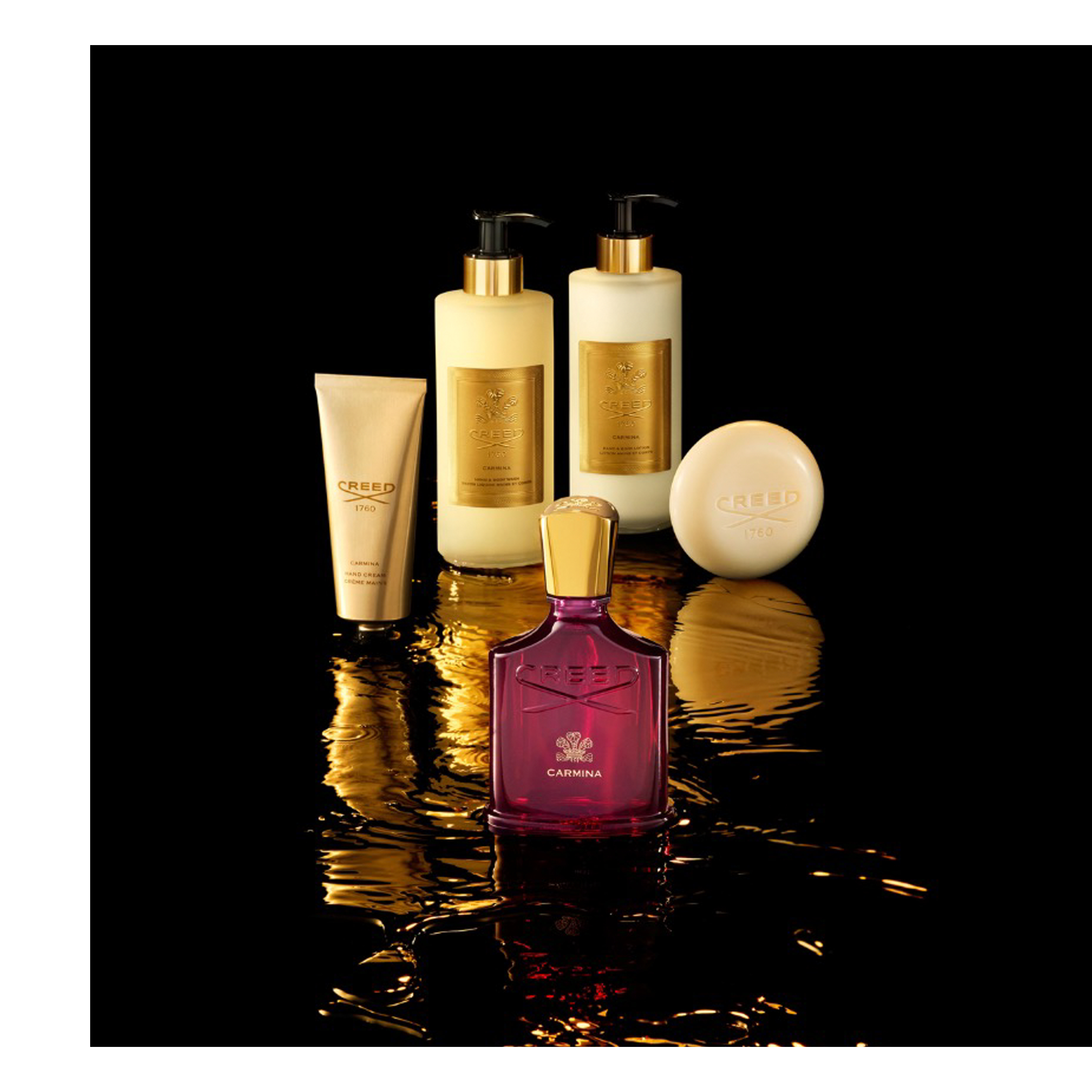 A luxurious collection of five Creed Carmina fragrance and body products is arranged on a glossy black surface with golden liquid reflections: a prominent dark red perfume bottle with a gold cap, a golden tube of hand cream, two tall golden pump bottles for hand and body wash and lotion, and a round cream-colored soap, bearing the visible text Creed 1760 Carmina, Creed 1760 Carmina Hand Cream, Creed 1760 Carmina Hand & Body Wash, Creed 1760 Carmina Hand & Body Lotion Lait de Corps, and Creed 1760.