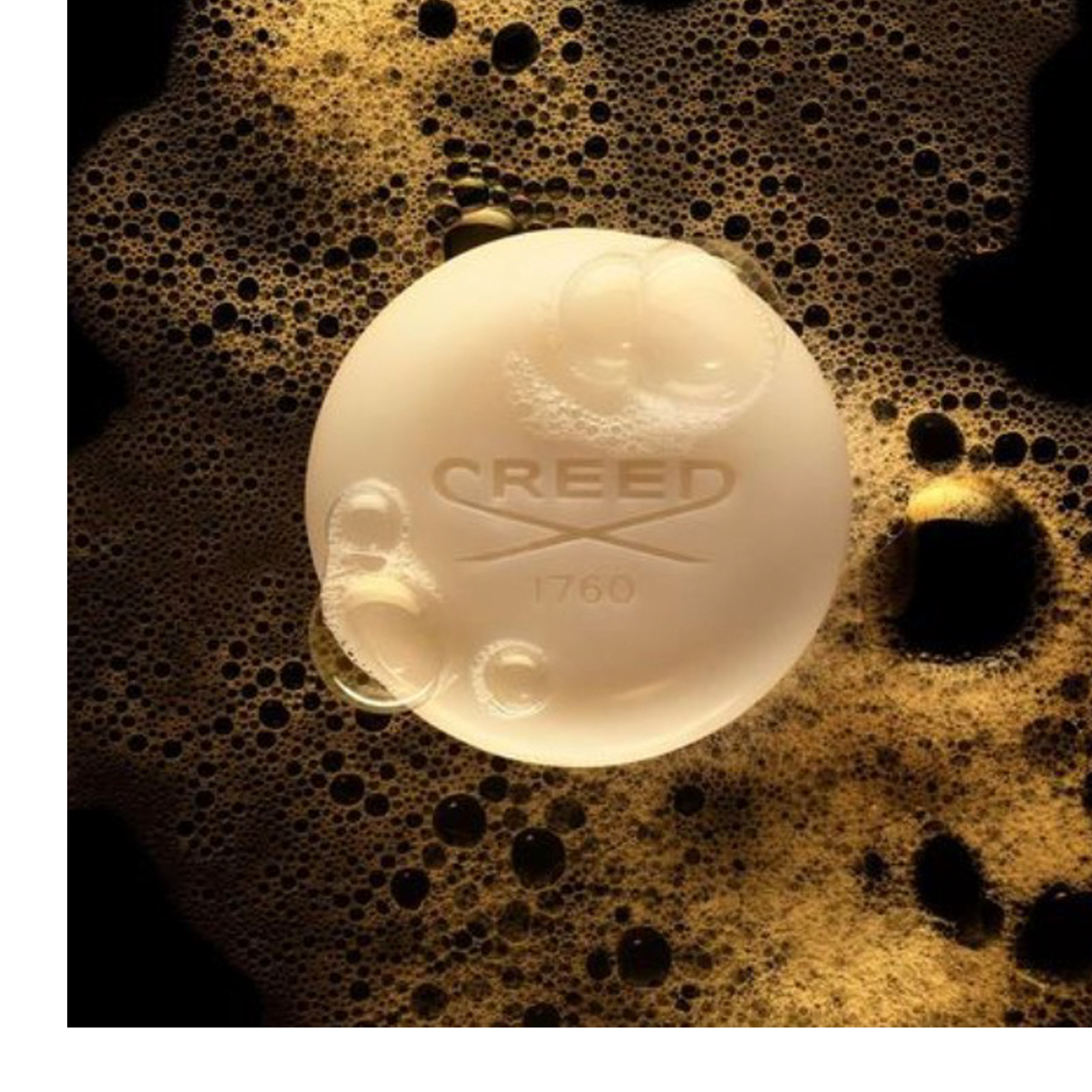  A round, creamy white soap bar with bubbles on its surface rests in golden-brown foam, displaying the embossed text CREED 1760. Image 1 - A round, creamy white soap bar with clear lather bubbles rests on a dark golden-brown bubbly foam, featuring the embossed text CREED 1760.