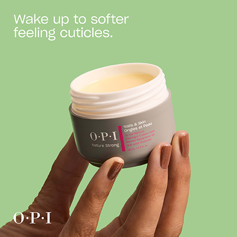 A hand with brown nail polish holds an open gray jar of OPI Nature Strong cuticle balm with yellowish contents, displaying text that reads Wake up to softer feeling cuticles, OPI, Nails & Skin, Ongles et Peau, To the Rescue, Overnight Cuticle Balm, Baume Reparateur de Nuit pour Cuticules, 20 mL / 0.7 fl. oz.