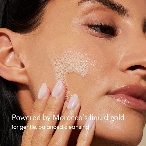 Image 1 - Close-up of a womans face with a hand gently applying a foamy cleanser to her cheek, with the text Powered by Moroccos liquid gold for gentle, balanced cleansing. Image 2 - A hand holds an izil The Argan Mild Balancing Cleanser bottle with orange liquid and a dark blue pump, while text next to it reads Deeply cleanses, Balances, Hydrates, and the bottle label also displays DEEPLY CLEANSE WITHOUT DRYING, BALANCE & SOOTHE, Argan Oil, Chamomile, Witch Hazel, NETTOYANT DOUX ÉQUILIBRANT À LARGAN, غسول متوازن خفيف بزيت الأركان, and ALL SKIN TYPES. Image 3 - An image displays a product redesign, showing two bottles of izil cleanser side-by-side with the text New Look Same formula you love, the old bottle on the left is labeled izil THE BASICS THE ARGAN MILD CLEANSER and the new bottle on the right is labeled izil THE ARGAN MILD BALANCING CLEANSER DEEPLY CLEANSES MITIGATES DRYING BALANCE & SOOTHES argan oil chamomile ALL SKIN TYPES.