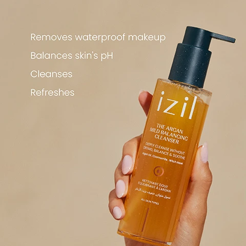 Image 1 - A hand holds a clear bottle of izil The Argan Mild Balancing Cleanser, a yellowish liquid with a dark pump, while text to the left lists its benefits: Removes waterproof makeup, Balances skins pH, Cleanses, Refreshes, and the bottle itself reads izil THE ARGAN MILD BALANCING CLEANSER DEEPLY CLEANSE WITHOUT DRYING, BALANCE & SOOTHE Argan Oil. Chamomile. Witch Hazel NETTOYANT DOUX ÉQUILIBRANT À LARGAN غسول متوازن خفيف بزيت الأركان ALL SKIN TYPES. Image 2 - A person uses a white cotton pad to remove makeup from their cheek, with text that reads Lifts stubborn makeup without stripping your skin.
