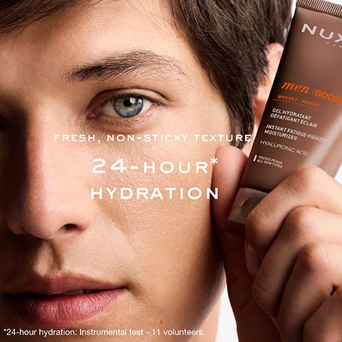 A close-up of a mans face, with a NUXE men [BOOST] skincare tube held beside it, shows the visible text NUXE PAR men [BOOST] BREVET - PATENT* GEL HYDRATANT DÉFATIGANT ÉCLAIR INSTANT FATIGUE-FIGHTER MOISTURIZER HYALURONIC ACID TOUTES PEAUX ALL SKIN TYPES FRESH, NON-STICKY TEXTURE 24-HOUR* HYDRATION *24-hour hydration: Instrumental test - 11 volunteers.