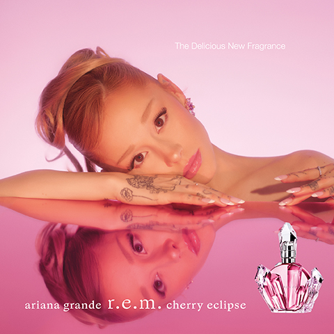 Ariana Grande leans on a reflective pink surface with her reflection visible below, a pink jewel-shaped perfume bottle to her right, with the text The Delicious New Fragrance and ariana grande r.e.m. cherry eclipse.